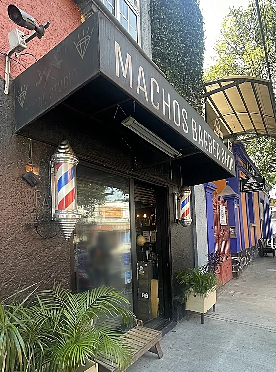 Entrada de la barbería con el toldo gris carbón que dice Machos Barber Shop, dos postes barberos, plantas y una banca de madera sobre la banqueta