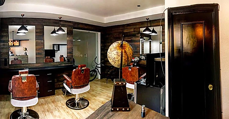 Interior de la barbería: tres sillones de cuero cognac frente a espejos, muro de tablones oscuros, lámparas colgantes, un globo terráqueo antiguo y una puerta de madera negra