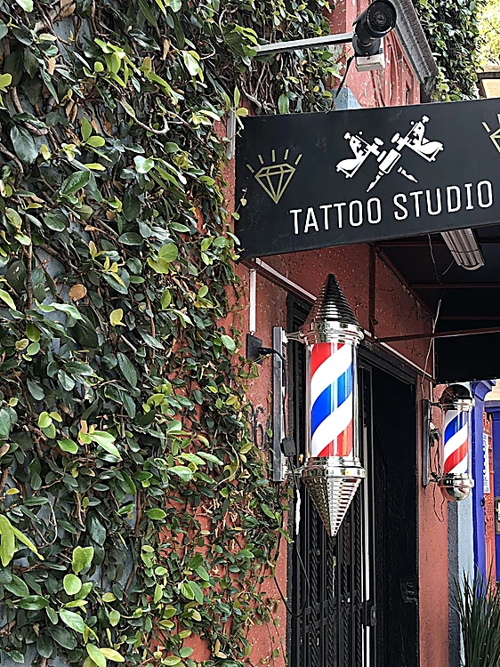 Muro cubierto de hiedra junto al letrero negro de Tattoo Studio con máquinas de tatuar y un diamante, y el poste barbero encendido